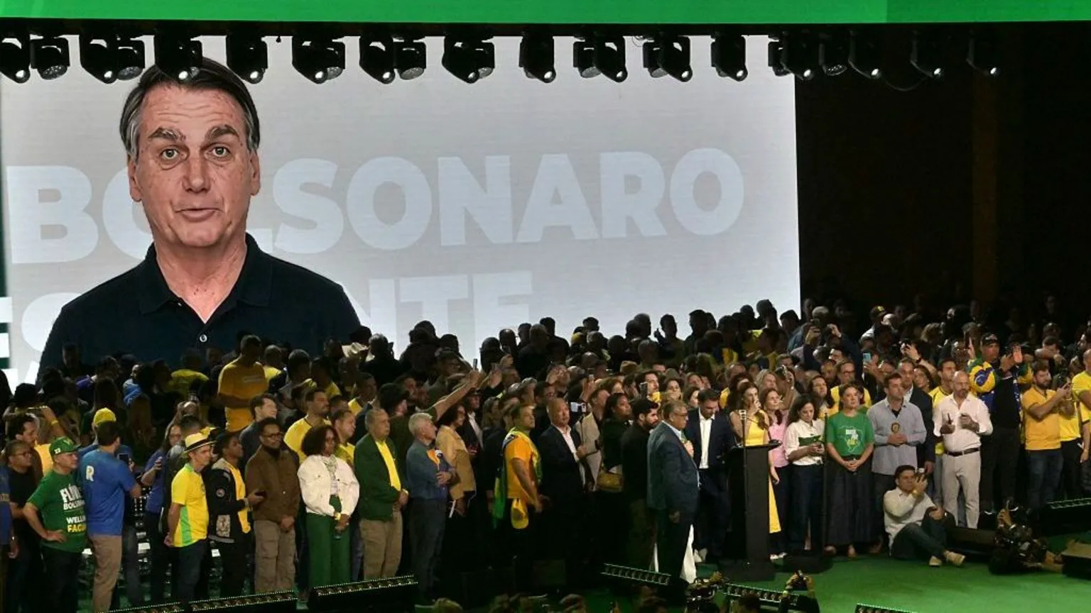 Nelson ALMEIDA / AFP via Getty Images An AI-generated video of former president Jair Bolsonaro is screened during the launch of the presidential candidacy of his son, Flávio Bolsonaro, at the Liberal Party convention, in São Paulo on 25 July, 2026. The image of Jair Bolsonaro, wearing a black polo shirt, appears against a grey and white background with the words "Bolsonaro presidente" emblazoned across it. In the foreground, Flávio Bolsonaro can be seen standing in front of a lectern. He is surr