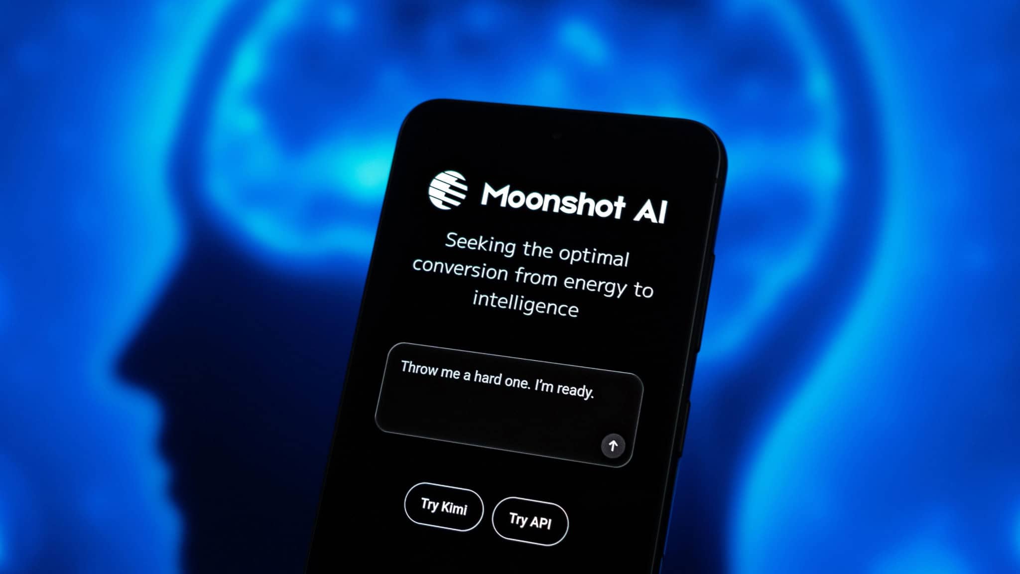Celular com Moonshot AI aberto; ao fundo, monitor exibe ilustração de cérebro em silhueta de cabeça humana